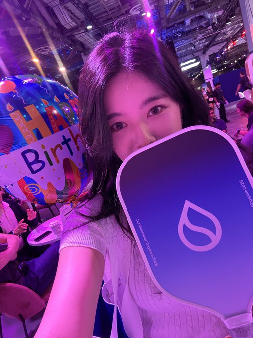 A young woman taking a selfie at what appears to be a birthday celebration, holding a paddle that partially covers her face, with colorful balloons and party lights behind her.