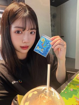 The image features a woman holding up a small blue card with a picture of a painting, possibly a Van Gogh painting, as she poses for the camera. The card has a straw sticking out of it. The background appears to be a dining table or countertop area where food and drinks are present.