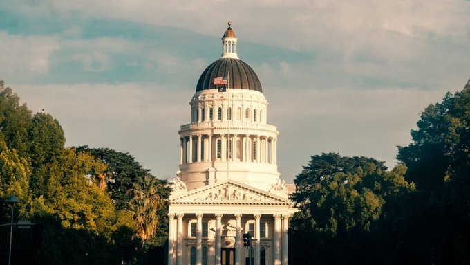 A stately government building with a prominent dome and classical columns, surrounded by trees under a partly cloudy sky.