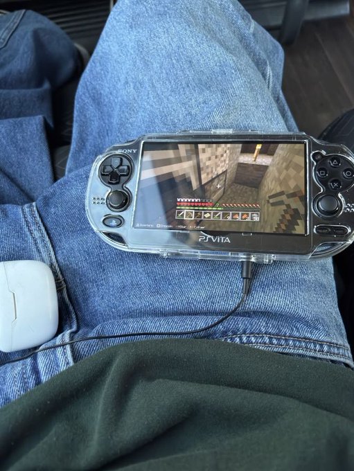 A Sony PlayStation Vita in a clear case rests on someone's lap, displaying Minecraft on its screen with a heart meter and inventory visible. The image relates to the post by showcasing the PS Vita—a handheld console with a loyal but niche fanbase—being actively enjoyed, illustrating the idea that dedicated owners remain committed to their device long after mainstream support faded.
