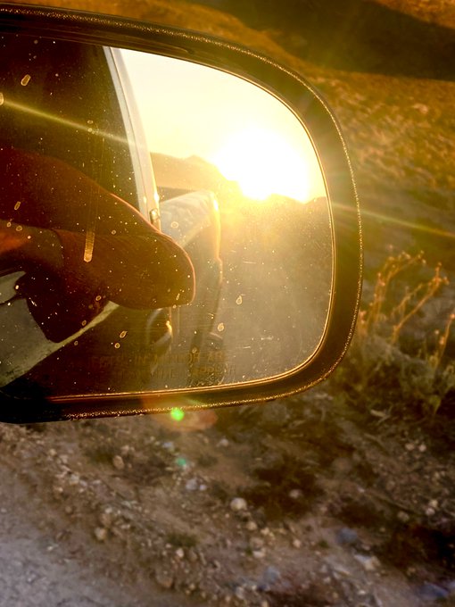 A driver's hand is reflected in a car's side mirror during a golden sunset, with sunlight flaring through the dusty glass.