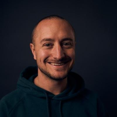 A headshot portrait shows a smiling man with short hair and a light beard, wearing a dark green hoodie against a dark background.