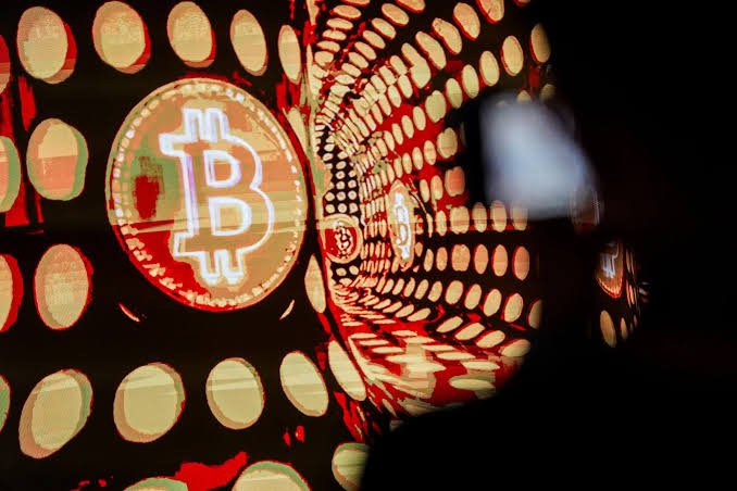 A glowing red Bitcoin logo projected onto a curved surface of dotted lights, with a silhouette of a person observing it in the foreground.