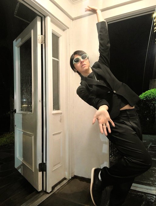 A person dressed in a black suit and sunglasses poses in a leaning stance near an open white doorway at night. The image has a dynamic, stylized aesthetic.