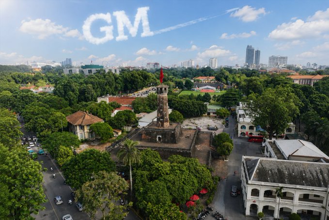 The user wants a concise caption for the image.
1.  **Analyze the image:** It's an aerial view of a city, likely Ho Chi Minh City given the "GM" text (likely standing for "Gia Miễn" or similar local slang/branding, but actually looks like "GM" in clouds). Wait, looking closely at the clouds, it spells "GM". The text in the prompt says "Buổi sáng vui vẻ nha ae ơii Mấy nữa lại được nhận PayX roàiii". This is Vietnamese.
2.  **Identify key elements:**
    *   **Sky:** Blue sky with white clouds. One cloud formation clearly spells "GM".
    *   **Foreground/Midground:** A historical-looking stone tower (looks like the Tower of the Citadel or similar structure, possibly the One Pillar Pagoda style but distinct, actually looks like the Tower of the Citadel of Saigon or similar historical