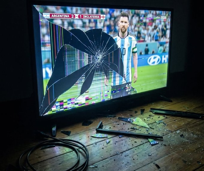 The image shows a television screen displaying a soccer match between Argentina and Inglaterra with a score of 2-1, featuring player Lionel Messi, while the screen is severely cracked and shattered, with debris scattered across the wooden floor.
