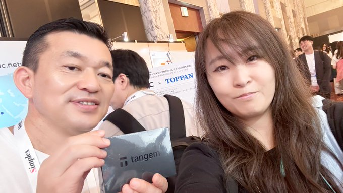 A selfie of a man and woman at what appears to be a conference, with the man holding a 'tangem' branded card, and a 'TOPPAN' banner visible in the background.
