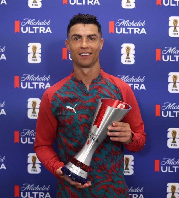 Cristiano Ronaldo smiling while holding a FIFA trophy at an event with Michelob Ultra branding in the background.
