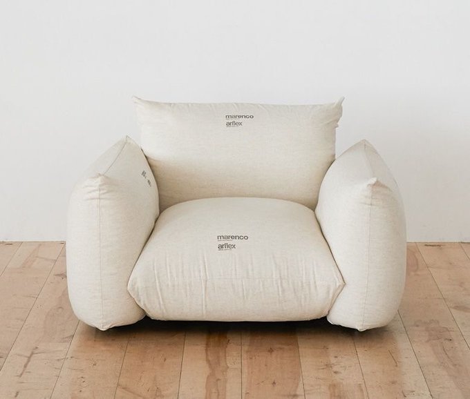 A cream-colored marenco arflex armchair and matching ottoman with a loose, puffy linen design sitting on a wooden floor against a white wall.