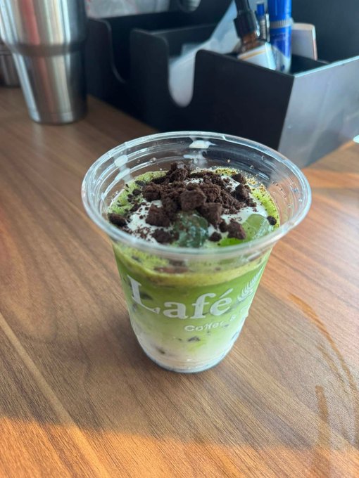 A close-up of a matcha coffee drink topped with crushed Oreo cookies and cream, served in a clear plastic cup with 'café' branding on a wooden table.