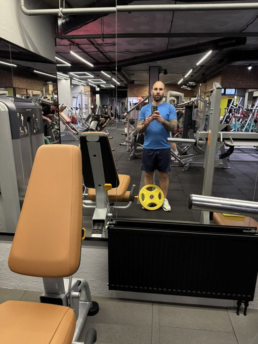 A man in a blue shirt and dark shorts taking a mirror selfie at a gym with weight machines, barbells, and weight plates visible around him.