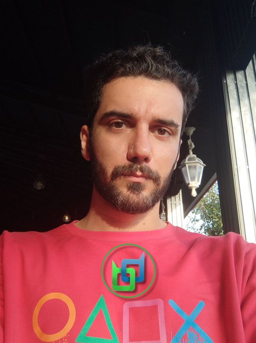 A bearded man takes a selfie wearing a red t-shirt featuring colorful PlayStation symbols (circle, triangle, square, X) under an outdoor covered area.
