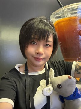A selfie of a young woman with short dark hair holding a large iced coffee drink, with a gray plush rabbit toy visible in the background.