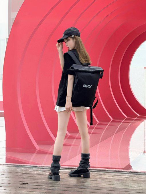 The image features a woman standing next to a red archway or tunnel-like structure. She has her back turned towards the camera while wearing a black backpack with "OKX" written on it, as well as a hat and boots. The background of the photo is blurred, but there are some buildings visible in the distance.