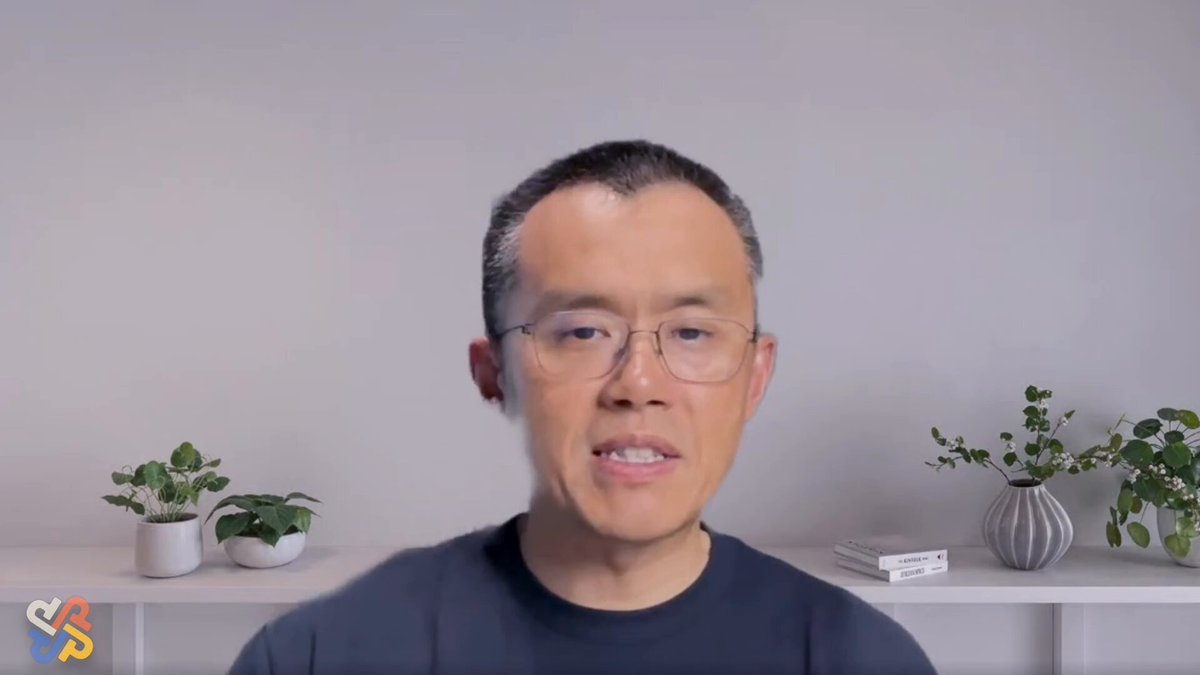 The image features a man wearing glasses, sitting at a desk with two potted plants behind him. The background of the image shows a white wall, which contrasts with the gray color of his shirt. There are also some books placed on the desk in front of him. The main focus of the image is the man's face and expression as he looks directly into the camera.