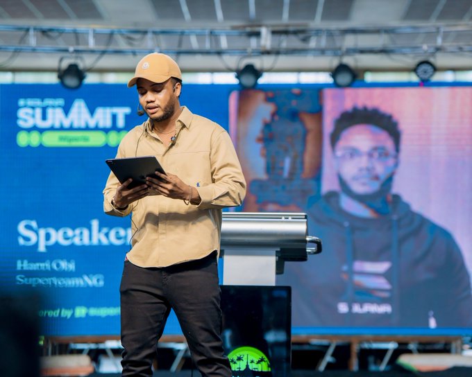 A man in a yellow shirt and cap speaks on stage while holding a tablet at the Solana Summit Nigeria event, with the screen behind him identifying him as "Harris Oli, SuperteamNG" under the "Speaker" banner. The image aligns with the post's reference to Solana Summit Nigeria as a flagship gathering for the SuperteamNG ecosystem in Nigeria.
