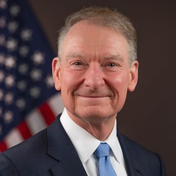An official portrait photograph of an older man in a dark suit and blue tie smiling, with an American flag visible in the background.