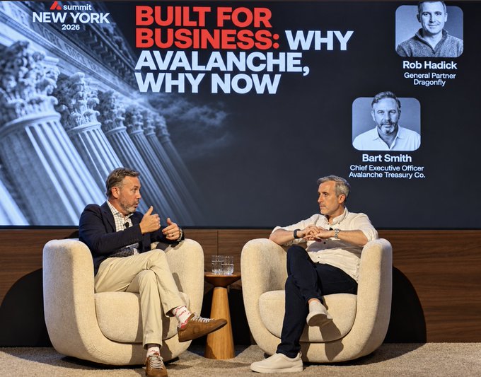 Two men sit in cream-colored armchairs on a stage at the Avalanche Summit New York 2026, with the screen behind them reading "BUILT FOR BUSINESS: WHY AVALANCHE, WHY NOW" and featuring photos of Rob Hadick (General Partner, Dragonfly) and Bart Smith (Chief Executive Officer, Avalanche Treasury Co.). The caption aligns with the on-stage discussion between the two executives, captured mid-conversation as Bart Smith explains why companies are hesitant to adopt blockchain technology despite pressure from their investors.
