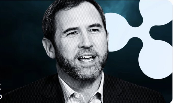 A black-and-white image of a bearded man in a suit speaking, with a large white Ripple (XRP) logo in the background.