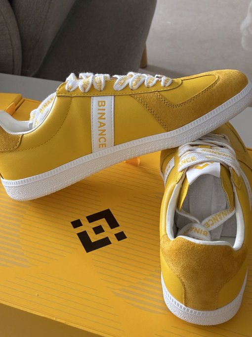 A pair of yellow sneakers with white laces and "BINANCE" branding on the side stripes, displayed on top of a yellow Binance shoe box featuring the black Binance logo.
