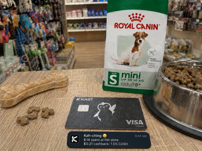 The image shows a table with two items - a bag of Royal Canin dog food and a Visa card. The table also has some treats for dogs placed nearby. A text overlay on the image reads, "Went to visit a friend today and couldn't show up empty-handed when there's a dog involved Picked up some treats on the way and paid with my KAST card. A few seconds later: kah-ching got my cashback." The post seems to be promoting a pet store or a pet care service, possibly offering rewards for customers who bring their pets in for purchases.