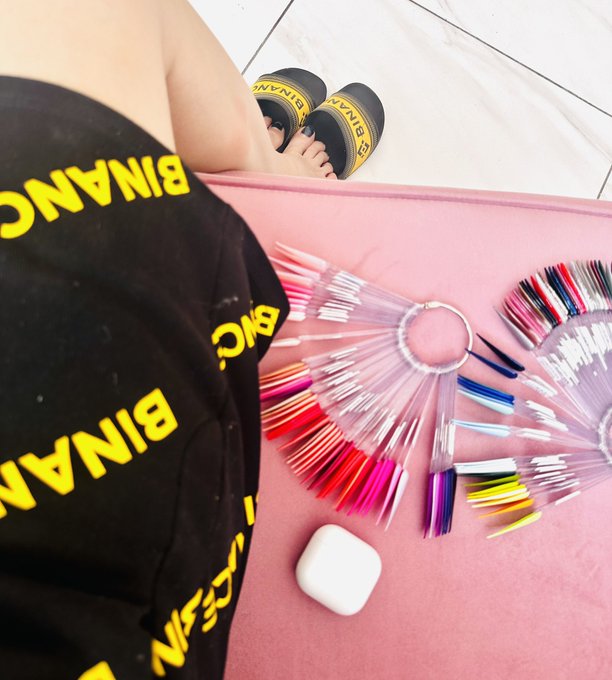 The image shows a person's feet wearing black sandals with yellow letters, sitting on a pink surface. The background features a pink wall with various nail polish bottles scattered around, suggesting that someone has been working on their nails recently or is preparing for a manicure session.