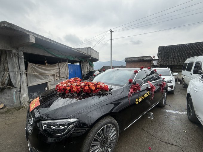 A black luxury car is decorated with red floral arrangements and ribbons for a wedding, parked outdoors in a rural area with a 'BINANCE' sticker visible on the door.