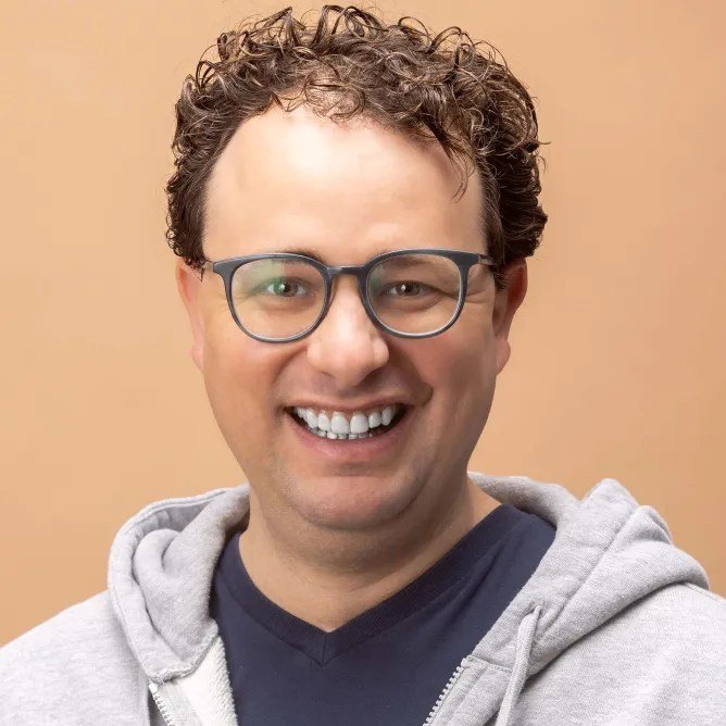A professional headshot of a smiling man with curly brown hair and glasses, wearing a gray hoodie over a navy blue shirt.
