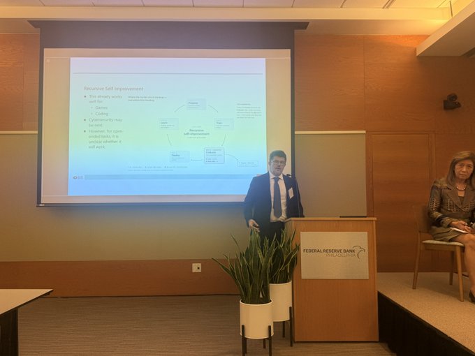 A presentation at the Federal Reserve Bank showing a man at a podium with a slide titled "Recursive Self Improvement" while a woman sits to the side.