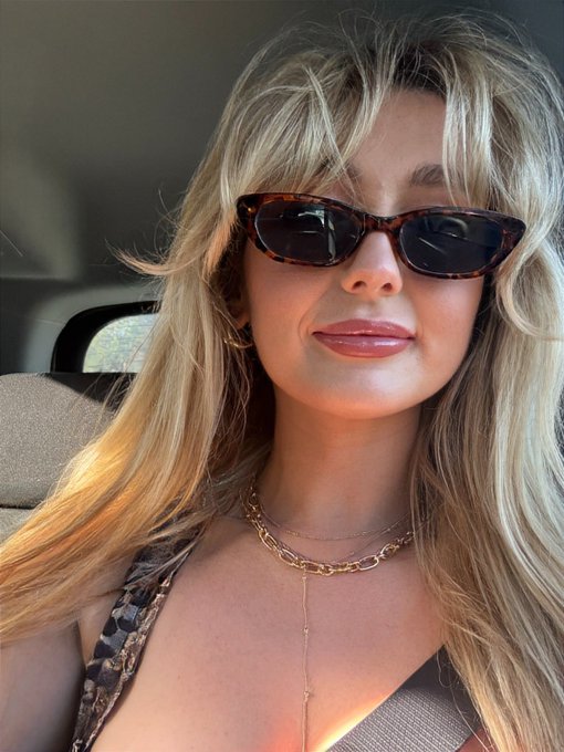 A blonde woman wearing tortoiseshell cat-eye sunglasses and layered gold necklaces takes a selfie in a car, with sunlight illuminating her hair.