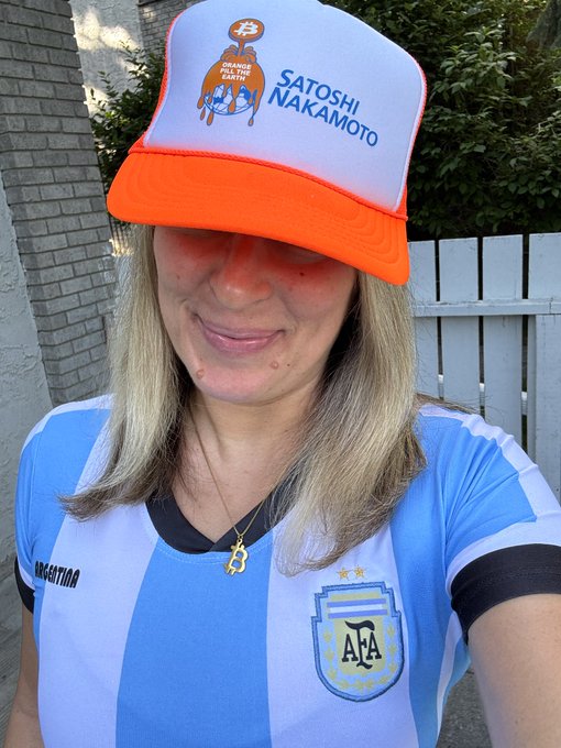 A woman wearing an Argentina national football team jersey and a white and orange trucker hat that reads 'SATOSHI NAKAMOTO' with an 'Orange Pill of the Earth' Bitcoin-themed logo.