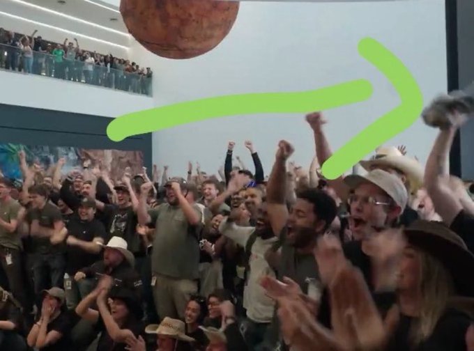 A crowd excitedly cheers with raised fists in what appears to be a SpaceX event or presentation, with a large spherical object (likely representing an asteroid) visible above them and an arrow highlighting it in the upper left of the image. The green arrow draws attention to the celestial body, aligning with the post's mention of spotting $ASTEROID in the SpaceX movie.