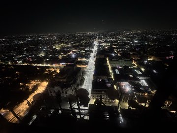 A nighttime aerial view of a long, straight city street lit up with traffic and streetlights, with a partial view of a camera equipment in the foreground.