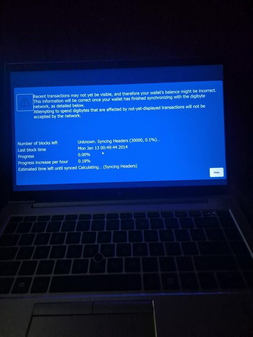 A laptop screen in a dark room displaying a cryptocurrency wallet sync error message, showing 'Number of blocks left: Unknown, Syncing Headers (30000, 0.1%)' and last block time 'Mon Jan 13 00:49:44 2014'.
