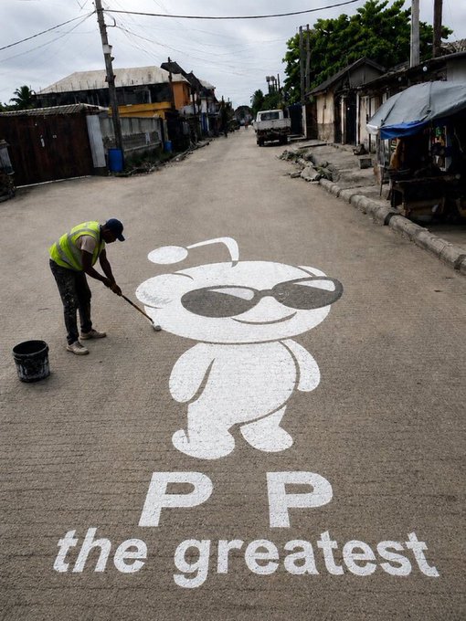 A worker in a high-visibility vest paints a large white logo on a street featuring a cartoon baby character wearing sunglasses alongside the text "P P the greatest," reflecting the post's playful appeal to "P P" for a blessing in the SHUFFLE game context.