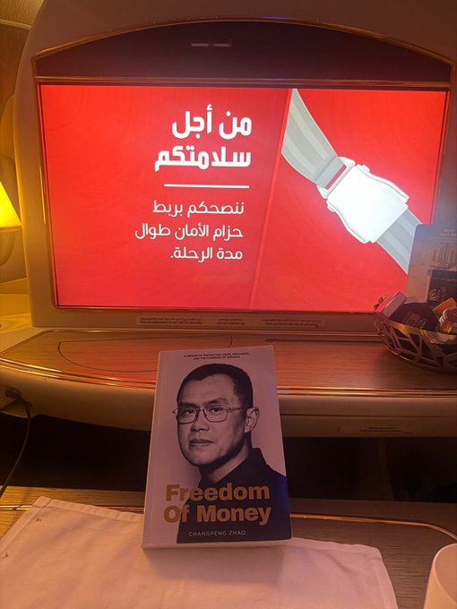 A book titled "Freedom of Money" by Changsong Zhao sits on an Emirates flight tray table in front of an in-flight entertainment screen displaying an Arabic safety announcement about fastening seatbelts, suggesting the traveler is en route to Astana, Kazakhstan.
