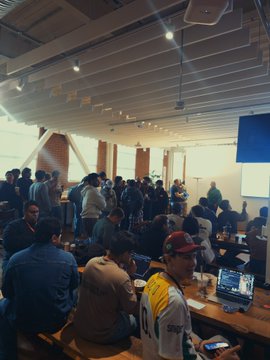A crowded room filled with developers and attendees seated at long tables working on laptops, with one person wearing a red cap and a yellow lanyard visible in the foreground. The packed standing-room-only atmosphere aligns with the post's description of a packed "grok bot hang" event.