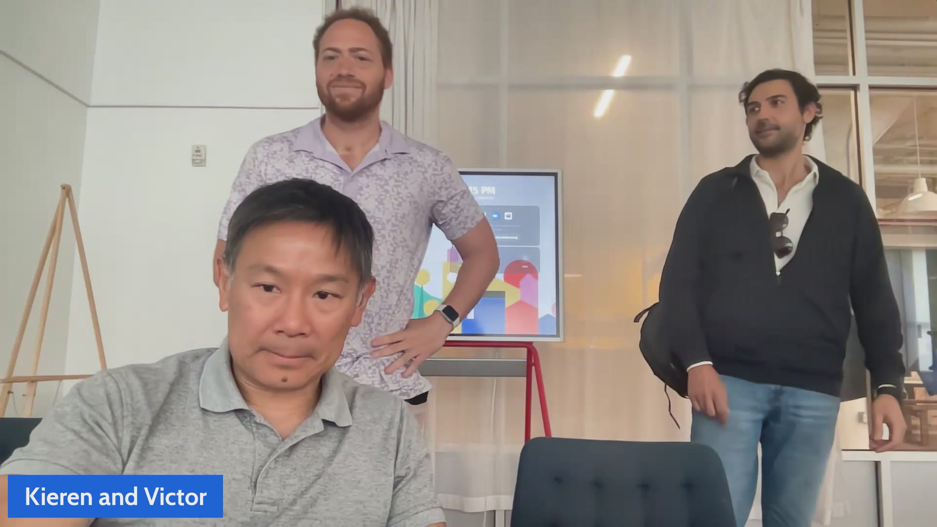 The image features three men standing next to each other in a room with white walls. One man is sitting at a desk, while another man stands behind him. The third man is standing directly in front of them. They appear to be engaged in conversation or observing something on a screen that is mounted on the wall.