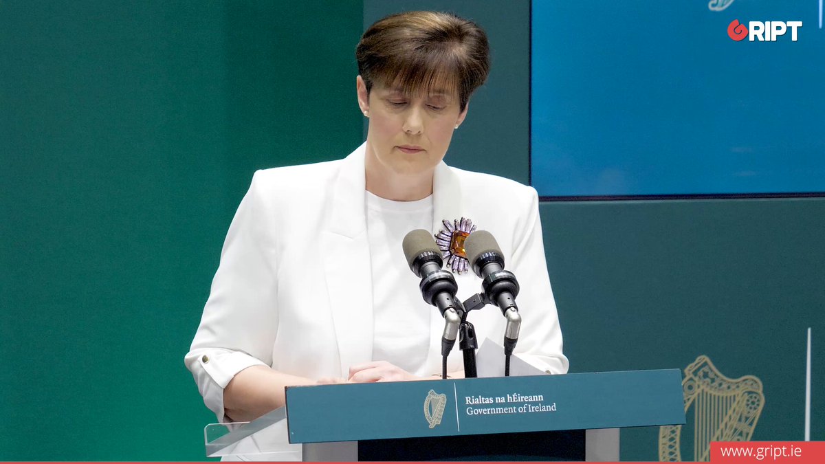 The image shows a woman standing at a podium, giving a speech or presentation. She appears to be wearing a white blazer and has her hands clasped together as she speaks into a microphone. The background of the image features a green wall with a blue sign on it that reads "RIPT".