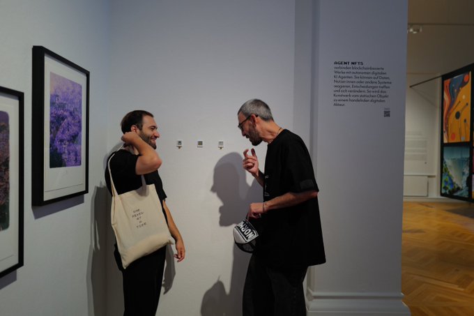 Two men converse in a museum gallery featuring framed digital artworks and an "AGENT NFTS" exhibition wall text discussing blockchain art, connecting to the caption's mention of "Agent NFTs in museums."