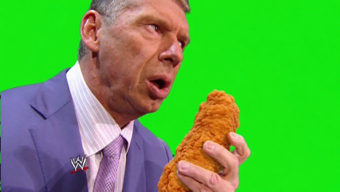 WWE commentator Vince McMahon in a blue suit and tie bites into a large piece of fried chicken against a green screen background.