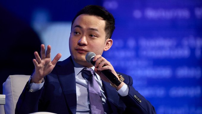 An Asian man in a dark suit and purple tie speaks into a microphone while gesturing with his other hand, appearing to participate in a panel discussion.