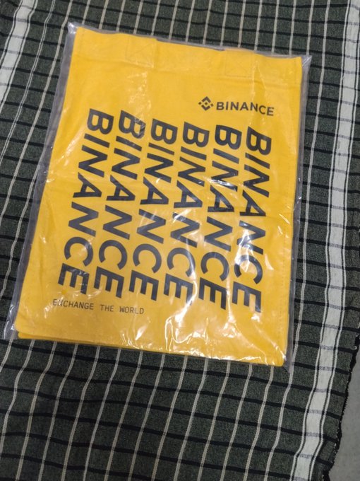 A yellow Binance merchandise t-shirt with 'BINANCE' printed repeatedly in black text and the tagline 'EXCHANGE THE WORLD' sits in clear packaging on a striped cloth.