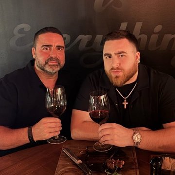 Two men sit at a restaurant table, each holding a glass of red wine, appearing relaxed as they pose for the photo; the caption suggests a humorous contrast between a casual dinner and the exaggerated "value" of their company, playing on the phrase "worth $300,000" likely referencing a viral meme about friendship and dining expenses.