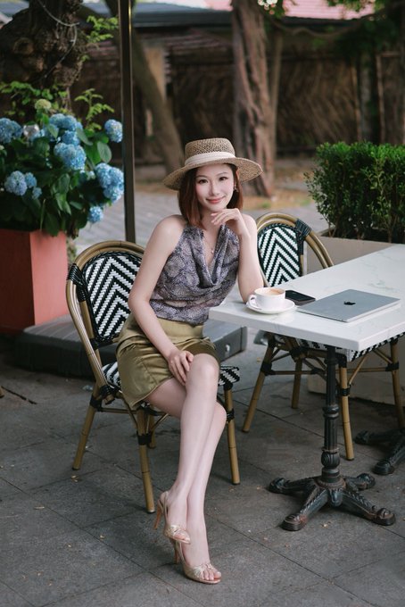 A woman in a straw hat, patterned sleeveless top, and olive skirt sits at an outdoor café table with a laptop and cappuccino, surrounded by lush greenery and blue hydrangeas. The relaxed, travel-friendly scene captures the jet-setting lifestyle of a crypto business developer hopping between global hubs like Hong Kong, Tokyo, and Singapore for Token2049.