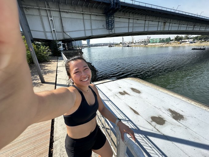 A young woman in athletic wear takes a selfie while jogging along a waterfront boardwalk, with a large bridge spanning the river behind her. The post says: "gm Belgrade! squeezing in a light jog before the @altitude x @lifiprotocol coffee afternoon!" The image captures the "light jog" part of her message, showing her in workout attire in what appears to be Belgrade, before heading to a coffee event.
