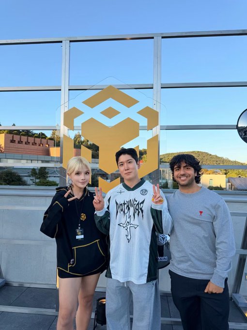 Three people pose outdoors in front of a large gold BNB Chain logo, with the central person wearing a shirt featuring stylized text. The image accompanies the post “Gmgn BNB Legends @BNBCHAIN @BNBCHAINZH,” linking the group photo to BNB Chain and its community.