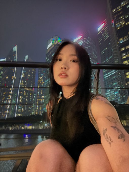 A young woman takes a selfie on a balcony overlooking Singapore's illuminated skyline, including the distinctive towers of the Marina Bay Sands complex, with "sg I am in u day 1 pre token :0" suggesting her excitement about arriving in Singapore. Her tattooed arm and casual pose give the photo a relaxed, personal travel diary vibe that matches the playful caption about being in Singapore on her first day.