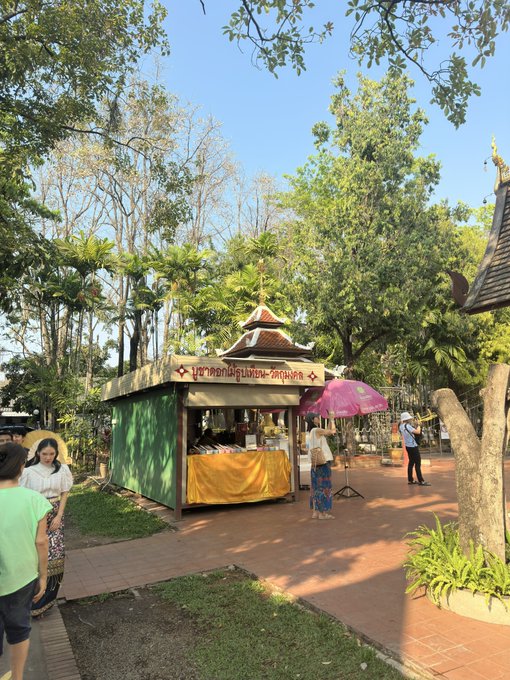 A small outdoor shop or kiosk with a green roof and yellow awning stands in a park-like setting, with a Thai sign reading "บูชาสังฆทาน-อัฐบริขาร" and people walking nearby.