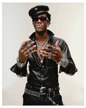 A photo of a stylish man wearing a black beret, sunglasses, a black leather shirt, multiple silver chains, large rings, and bracelets, posing with his hands up.